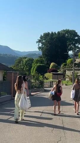 The video shows four young women walking down a paved road on a sunny day. They are all carrying bags, presumably heading somewhere for leisure. The woman closest to the camera is wearing a light-colored bikini top and loose pants. Behind her, one woman is wearing a black dress, another a patterned dress, and the last one, who is partially obscured, wears a light blue swimsuit. The background reveals a landscape of rolling hills, houses, and lush greenery. The sky is clear and blue. The women are walking away from the camera.