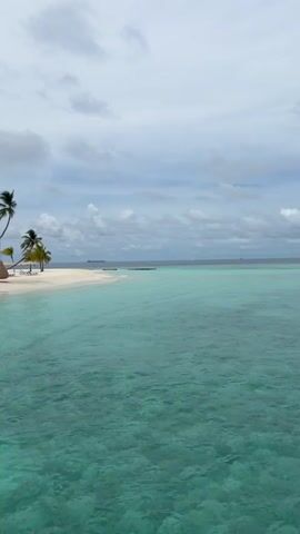 The video shows a slow pan across a tropical beach scene. The foreground is dominated by crystal clear turquoise water. In the middle ground, a white sand beach is lined with palm trees. Beach umbrellas and lounge chairs are visible, as are several thatched-roof structures, suggesting a resort. In the background, the sky is overcast with scattered clouds. There are no people clearly visible in the frame. The overall impression is one of a serene, idyllic island vacation setting.