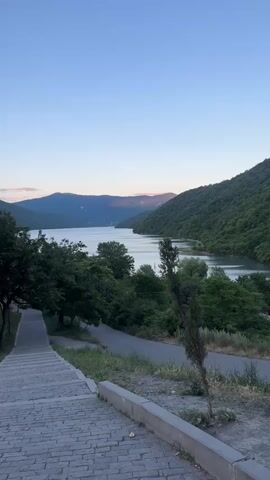 The video pans across a serene lake surrounded by lush green hills. In the foreground, a stone staircase leads down towards a paved path. The sky is a clear, pale blue, indicating either dawn or dusk. Sunlight casts a soft glow on the distant hills. The water of the lake is calm, with subtle ripples reflecting the surrounding landscape. Dense foliage covers the slopes of the hills, creating a sense of natural beauty and tranquility. There are no people or any noticeable activity other than the slow movement of the camera.