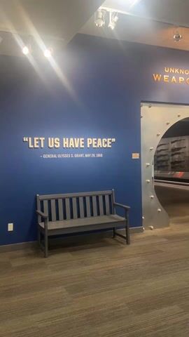 The camera slowly pans to the right, revealing a museum exhibit entitled "Unknown Soldiers Weapons Vault." The entrance is designed to look like a vault door made of polished silver metal, with a barred gate further inside. The door is open, revealing a display of weapons inside. To the left of the entrance is a blue wall with the quote "Let us have peace" - General Ulysses S. Grant, May 29, 1868. A gray wooden bench sits in front of the wall. The floor is covered in brown and gray carpet. The lighting is bright, with spotlights illuminating the wall and entrance.