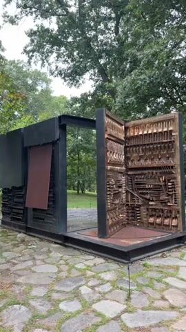 The video shows a large outdoor sculpture made of metal and wood. The sculpture resembles an open book or cabinet with many compartments filled with various objects, primarily tools and hardware. The sculpture is set on a stone pathway, surrounded by trees and grass. The camera slowly pans around the sculpture, offering different angles and perspectives. The weather appears to be overcast, with soft, diffused light. There are no people visible in the video.