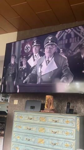 The video shows a television screen displaying a black and white scene of a man in a Nazi uniform speaking at a podium. The background includes other uniformed figures and Nazi flags. The TV is mounted on a wall above a light blue dresser with gold accents. A black wireless router and a decorative wooden sculpture are placed on top of the dresser, along with a black remote control. The environment suggests an indoor setting with a wooden ceiling.