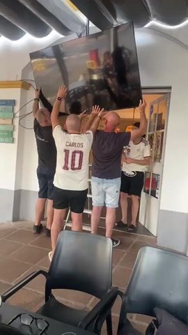 Four men are carrying a large flat-screen television. One man on the left is wearing a black t-shirt and dark shorts. The second man from the left is wearing a white soccer jersey with the name "CARLOS" and the number "10" on the back, and dark shorts. The third man from the left is wearing a dark t-shirt and light blue denim shorts. The fourth man on the right is wearing a white soccer jersey with black trim and dark shorts. They are all holding the TV above their heads. The TV is angled upwards. They appear to be in an outdoor or semi-outdoor area with a tiled floor. There are chairs in the foreground. Above them, there is a structure with dark, overlapping panels that appear to be part of a roof or awning. To the right, there is a doorway or opening with glass. The lighting suggests it is daytime.