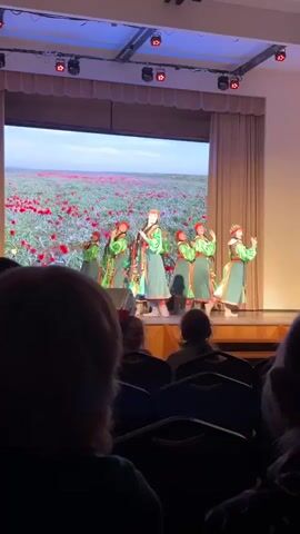 On a theater stage, approximately a dozen young women are performing a traditional dance. They are dressed in bright green and red folk costumes. Behind them, a large screen displays a field of red tulips. The dancers move in unison, their arms flowing as they turn and circle. The lighting is bright, highlighting the colors of their costumes. The scene is viewed from the audience perspective. Dark figures of people sitting in rows of chairs are seen in the foreground.