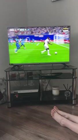 The video shows a television screen displaying a soccer match. The score displayed on the screen indicates "ENG 1" and "COD 1". The game appears to be a live broadcast, with a timer showing "51:41". The stadium is filled with spectators, and advertisements for "Budweiser" are visible around the field. On the bottom shelf of the TV stand, a DVD player, a scented candle, and a reed diffuser are present. In the foreground, a person's bare feet are visible, suggesting they are watching the game from a relaxed position on the floor. The room has wooden flooring and a grey wall. The overall setting appears to be a living room in a residential area. The game seems to be in progress, with players actively engaged on the field. There is no explicit information about the time of day, weather, or specific emotions displayed by the people. The location context provided mentions Vyzhnytsia, Ukraine.