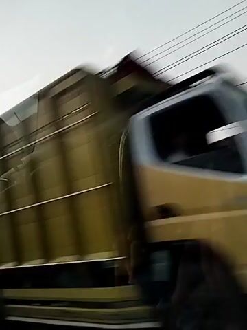 The video shows a view from inside a vehicle traveling on a road, likely in a rural or semi-urban area. Several utility poles with numerous wires are visible, stretching across the frame. The landscape consists of dry grass, bushes, and some trees. In the distance, there appear to be some buildings. At various points, other vehicles pass by. Specifically, a black car is seen driving past. Later, a silver truck with a covered cargo bed is also captured as it drives by. The sky is overcast, suggesting either early morning or late afternoon. The overall impression is of a roadside scene captured during a journey.