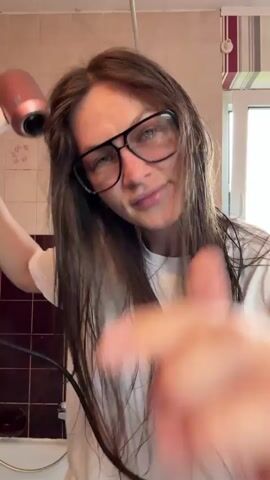 A woman with long, dark hair and glasses is drying her hair with a pink hairdryer in a bathroom. She is wearing a white t-shirt. The bathroom has tiled walls.