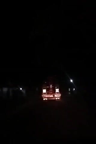 At night, a dimly lit truck with its rear lights on is seen from behind on a road. Other faint lights are visible in the distance. The overall scene is dark and the truck is the primary focus.