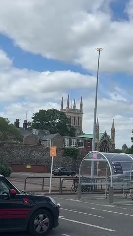 The video presents a static, eye-level, daytime shot of an urban landscape under a cloudy blue sky. In the immediate lower left foreground, the front quarter of a black Mini Cooper with a red stripe on its side is visible. To the right, a silver, tall street lamp with a circular light fixture stands next to a modern, arch-shaped, transparent shelter, possibly for shopping trolleys, which features a clear roof and metal frame. Two rectangular signs with a shopping cart icon are visible near the shelter, one orange and one pink. Further back, a dark-colored sports car is parked in what appears to be a parking lot. Behind the parking area, a low stone wall supports a row of buildings with traditional architecture, including a building with a green roof. A large, verdant tree is situated behind these buildings. Dominating the background is a large, elaborate stone church tower with multiple pointed spires. Throughout the video, the distinct sound of church bells ringing is audible.