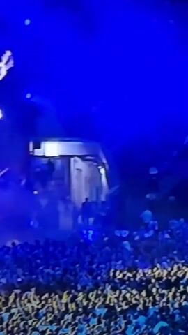 A concert crowd is visible in the foreground, looking up towards a stage. The stage is illuminated by intense blue lighting. A large silhouette of an electric guitar hangs from above. Some stage equipment, including lighting rigs and possibly speakers, is visible. The crowd appears to be engaged and watching the performance.