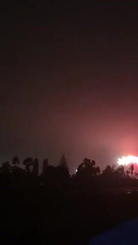 The video shows a nighttime outdoor scene with fireworks being set off. The fireworks are visible as bursts of light in the dark sky. Silhouetted trees and foliage form a dark line across the lower part of the frame, with a dark, out-of-focus foreground that appears to be a surface. The sky is dark, with some haze or smoke from the fireworks. The fireworks themselves are colorful, with white, pink, and possibly other colors visible. The scene suggests a celebration or event taking place after dark.