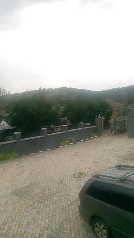 This is a still image with a timestamp that suggests it may be part of a video. The scene is an outdoor environment, likely a paved area next to a row of buildings or structures. The image is oriented vertically, with the top of the frame appearing on the left side and the bottom on the right.

The primary subjects in the image include a dark-colored vehicle, possibly an SUV or minivan, parked on a grey, interlocking brick paved surface. Beyond the paved area, there is a section with several identical dark-colored structures, each with a decorative metal railing or fence. These structures are set against a backdrop of lush green trees that cover a hillside. The trees are dense and extend upwards, suggesting a natural or park-like setting behind the man-made structures. The sky is overcast, indicating a cloudy day.

There is no clear activity taking place; the scene appears to be a static view of the location. The notable details include the paved surface, the modern design of the railings, and the abundant greenery. The location context provided is "Ire, Nigeria," which helps to place the scene in West Africa. There is no visible text within the image itself. The overall mood is neutral, with the overcast sky contributing to a somewhat subdued atmosphere.
