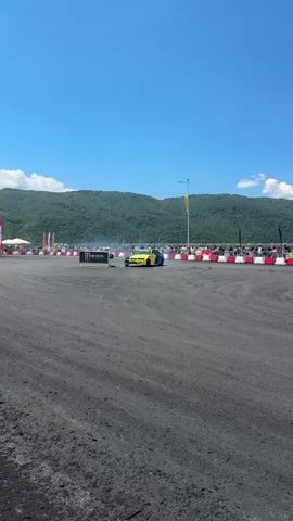 The video shows a car drifting event on a sunny day. Several cars are visible, some with colorful liveries. The cars are performing controlled slides around a track. In the background, there are spectators behind barriers and a scenic view of mountains. The track surface appears to be asphalt or dirt. There is a significant amount of tire smoke being produced by the cars as they drift. One car is predominantly blue and yellow with "FALKEN" written on it. Another car is white with some racing decals. A yellow and blue car is also visible. The sky is clear blue with a few white clouds. Tall banners are placed along the sides of the track, some with logos such as "Shell Power" and "Monster Energy".
