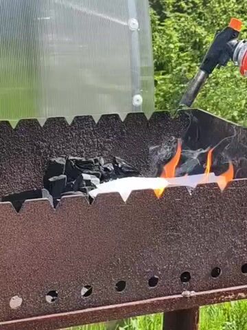 The video shows a metal grill being lit with a torch. The grill is filled with charcoal. The torch is black and orange. There is a clear, corrugated plastic panel behind the grill. Green trees are visible in the background. The grill is placed on a green lawn. Smoke and flames are emanating from the grill.
