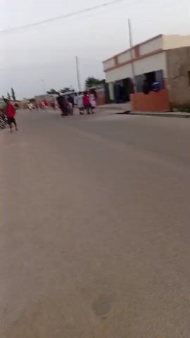 A group of people are walking or running down a paved road. They are in a line and appear to be moving in the same direction. Some individuals are wearing red shirts, while others are wearing white and darker clothing. In the background, there are buildings, possibly shops, with awnings and windows. The sky is overcast. There is a street light visible on the left side of the road. The overall scene suggests an outdoor activity or gathering in a town or village.