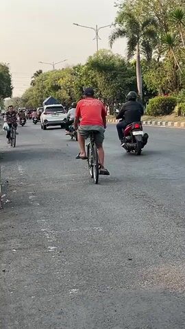 A busy street scene with multiple people and vehicles. In the foreground, a person rides a bicycle on the right side of the road, facing away from the camera. To the left, another person rides a bicycle towards the camera, also on the right side of the road. Further down the road, several motorcycles and cars are visible, along with more trees and streetlights lining the sides of the street. The overall atmosphere suggests a typical urban environment with moderate traffic.