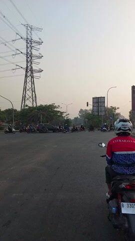 This is a first-person perspective video filmed from the back of a motorcycle in Pasir Jaya, Indonesia. The scene captures a procession of motorcycles and cars traveling along a paved road. The rider in the foreground wears a red jacket with "CITRARAYASC" written on the back and a white helmet with blue accents. Behind them, a line of motorcycles and a few cars are visible. In the distance, tall power line towers are prominent, suggesting an industrial or semi-urban setting. The sky is overcast and hazy, indicating either early morning, late afternoon, or possibly smog. The overall atmosphere is one of everyday commuting or travel. Visible text includes the license plate "B 3305 CDL" on the motorcycle in the foreground and signage in the background that is too blurry to read clearly.
