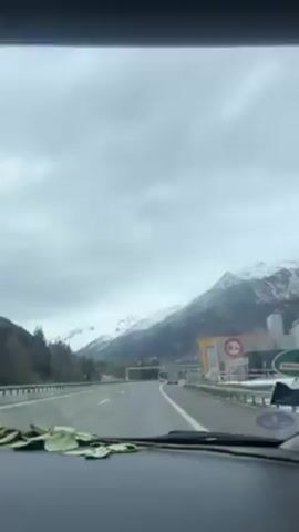 The video shows the interior of a car traveling on a highway against a backdrop of mountains partially covered in snow. The sky is overcast. Inside the car, the camera's viewpoint shifts to show the person in the passenger seat drinking from a red can of Coca-Cola.  After taking a sip, the person looks up and gives a slight smile.  The person is wearing a black Adidas hoodie.  The surroundings outside the car window are blurry as the car is in motion.