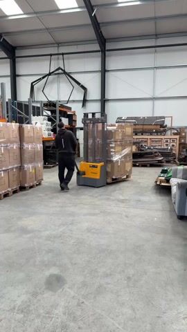 A man in a black hoodie and black pants is operating a yellow Jungheinrich electric pallet jack in a warehouse. He is pulling a load of boxes wrapped in clear plastic. To his left, there are stacks of boxes and a rack of equipment. To his right, there are more boxed items, some palletized, and a grey couch. The warehouse has a concrete floor, grey walls, and a black metal frame. There is a geometric metal structure hanging from the ceiling.