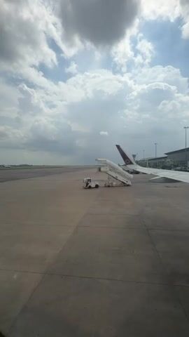 A tug is attached to a set of mobile airstairs that are positioned next to a large airplane. The airplane has a dark purple tail fin with a gold emblem. The airstairs are white and have a covered walkway. The tug is white and has a single operator visible inside. The tug is connected to the airstairs by a tow bar. The scene takes place on an airport tarmac under a cloudy sky. In the distance, a runway stretches out and other airport buildings and equipment are visible.
