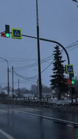 The video shows a road intersection in what appears to be a rural or suburban area on a gloomy, overcast day. The road is wet, and there are patches of snow along the side. Two sets of traffic lights are visible, both displaying pedestrian crossing signals. On the left, the light shows a red pedestrian signal and a green traffic signal. On the right, the traffic light is counting down from 13 seconds for pedestrians to cross, with both a red pedestrian signal and a green traffic signal illuminated. Above both sets of lights are pedestrian crossing signs. Utility poles and wires run along the side of the road, and a tall communication tower is visible in the background. A pine tree stands near the right set of traffic lights. In the distance, there are houses and other buildings, partially obscured by trees. Traffic can be seen in the distance as well.