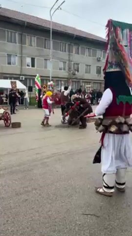 The video shows a group of people dressed in traditional Bulgarian Kouker costumes performing in a public square. The background includes a light gray, multi-story building, tents, and other people observing the performance. The camera focuses on one Kouker in particular, whose costume consists of a tall, colorful headdress, a red vest over a white tunic, and several large bells attached to the waist. In the center, several Koukers are wrestling on the ground, enacting a traditional performance. The overall setting is festive, with a cultural event taking place in an urban environment.