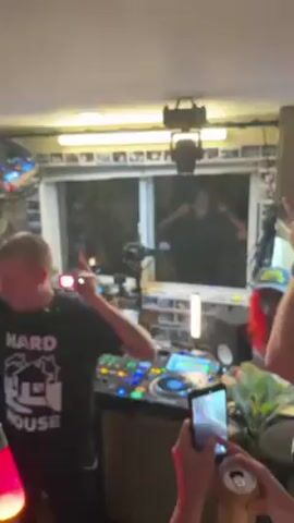 The video shows a DJ performing in what appears to be a small, crowded room.  The DJ, wearing a black "Hard House" t-shirt, stands behind a large set of DJ equipment. People are dancing and filming with their phones.  A lava lamp glows in the foreground. Behind the DJ, a window is visible, and someone is dancing outside. The room is cluttered, with various items and decorations visible.  Other people are seen in the background of the room, with a door and a television visible in the distance.