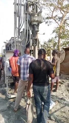 The video shows a group of men standing around a large drilling machine on a muddy, possibly construction, site. The machine is tall and has many pipes and hydraulic components. It is drilling into the ground. The men are watching the drilling process. Some are wearing casual clothes such as jeans and t-shirts while others are wearing vests and head coverings. There is a tree visible in the background.