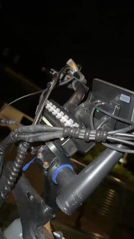 The video shows a close-up view of an electric bicycle at night. The camera pans down from the handlebars, which are cluttered with cables and a display screen, to the front wheel and fender. The bicycle frame is gray and black, with a battery pack mounted on it. There is a small bag attached to the frame. The background is dark, with glimpses of cars and a fence visible. The scene is illuminated by streetlights.