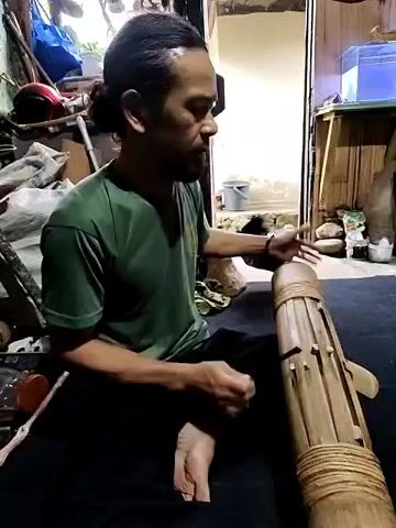 A barefoot, tan-skinned man with dark hair pulled back in a bun sits cross-legged on a dark surface. He is wearing a dark green t-shirt and dark pants. He plays a long, light-brown bamboo instrument that sits in front of him. He taps the side of the instrument with his left hand and strikes the top with a wooden stick in his right.  A room cluttered with objects is behind him. A blue tote, a helmet, an unidentifiable machine, and wooden planks are visible. A doorway is to the right of the man, providing a view to an outdoor scene. There is a container and some rocks outside.