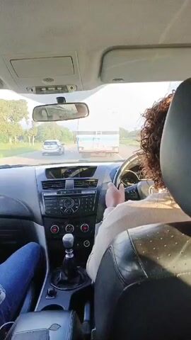 From the interior of a car, the video shows a road scene with a driver and a passenger. The driver has curly hair and is wearing a light-colored sweater. The passenger is wearing blue jeans. The car is stopped on the road, waiting for pedestrians to cross. In front of the car, there is a silver car, a blue and white truck, and two pedestrians walking across the street. The road is paved and there are trees on either side. The sky is overcast with some visible blue patches. The car's dashboard is visible, featuring the steering wheel, gear shift, radio, and air conditioning controls.