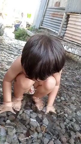 A child, wearing only a patterned diaper, crouches in shallow, rocky water. The child then stands and walks, splashing the water. In the background, two people are visible, partially obscured by foliage and a corrugated metal structure. The environment appears to be outdoors, possibly a stream or riverbed.