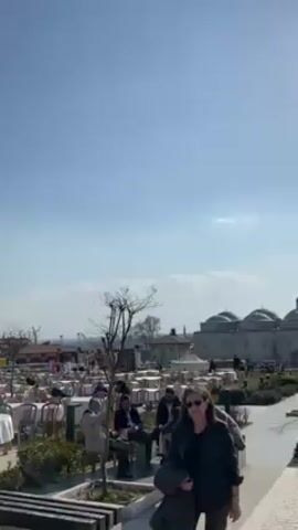 The video shows an outdoor scene with a large number of tables and chairs set up, possibly for an event or restaurant seating. In the background, there is a grand mosque with tall minarets. People are seen walking around and sitting at the tables. The sky is blue and mostly clear, indicating a sunny day. The camera pans to show the full view of the mosque and the outdoor seating area.