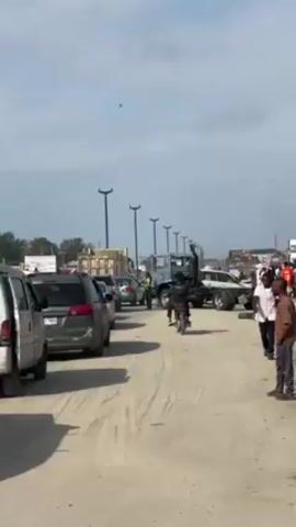 The video shows a traffic jam on a sandy road, possibly a beach access road or an unpaved street, with several cars lined up. A motorcycle navigates between the cars, while a truck stands nearby. There are people standing on the side of the road. The sky is overcast. There are streetlights lining the road.