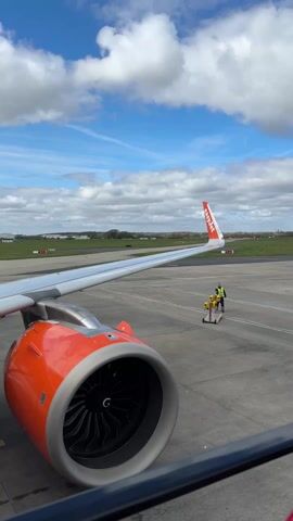The video shows an EasyJet airplane wing and engine against a backdrop of an airport runway on a partially cloudy day. An airport worker in a yellow safety vest is visible in the distance, pulling a small yellow and silver cart with two yellow cylinders on it. The cart is moving towards the right of the frame on the runway. The EasyJet tail fin is orange and white with the airline's name on it. The engine is orange and silver. In the background, there is green grass and some buildings under a sky with a mix of blue and scattered white clouds.
