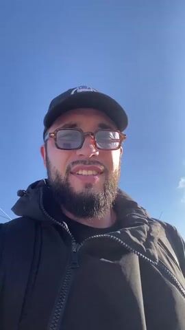 A man is recording a selfie video outdoors under a clear blue sky. He is wearing a black baseball cap, a black jacket, and glasses with thick, brown frames. He has a beard and is smiling at the camera. The sun is shining brightly, casting light on his face and the sky. There are a few faint clouds visible in the background. The man appears to be speaking, but his words are not transcribed. The video is shot from a slightly low angle, looking up at the man.