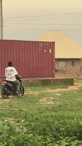 A young man rides a black motorcycle on a grassy patch. He wears a white t-shirt and patterned pants. Beside him is a large, red shipping container and a tan-colored building with a pyramid roof. The area appears to be a rural or semi-rural environment.