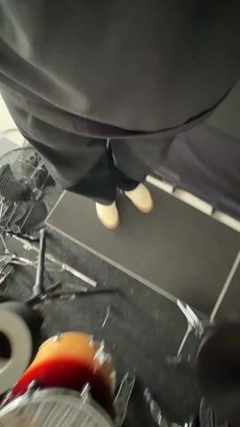 From an overhead perspective, the video shows a person standing on a black platform surrounded by a drum set and various musical equipment. The person is wearing black clothing and beige shoes. The drum set has a distinct orange and yellow ombre finish. Various cymbals and drum hardware are visible around the drums. The environment includes a fan, cables, and other music-related items scattered around. At one point the person makes a 'rock on' hand gesture.