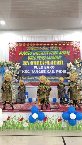 A group of five young children, dressed in elaborate gold and green costumes with ornate headpieces, perform a dance on a stage. The children are arranged in a staggered formation on the raised platform. Behind them, a large banner reads "SELAMAT dan SUKSES AJANG KREATIFITAS ANAK DAN PERPISAHAN RA DARUSSA'ADAH PULO BARO KEC. TANGSE KAB. PIDIE." The stage is decorated with garlands of white flowers and blue and white balloons. The floor in front of the stage is adorned with green paper grass and more blue and white balloons. The overall environment appears to be an indoor event space with a white ceiling and artificial lighting. The children are moving in unison, their hands raised.