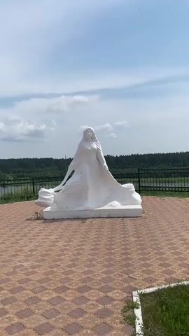 A white sculpture of a woman in a flowing dress stands on a tiled platform. The sculpture is a full-body depiction with the woman facing forward, her arms at her sides, and her dress dramatically fanned out around her. Behind the sculpture, a black metal fence separates the platform from a grassy area and a body of water, likely a river, with a line of trees on the opposite bank. The sky above is blue with scattered white clouds. The video appears to be filmed from a slightly low angle, looking up at the sculpture. The lighting suggests it is daytime, likely a bright, sunny day. The overall scene is peaceful and scenic.