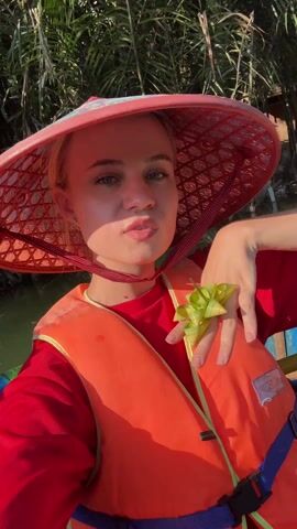 A fair-skinned young woman with blonde hair is taking a selfie. She is wearing a red shirt and an orange life vest with a blue strap. A large, red, conical Asian hat with a floral patterned brim is on her head, held in place by a red strap. She has a small, green woven flower in her hand, which she is resting against her cheek. She looks directly at the camera, then glances off to the side. The woman is seated in a colorful boat on a river, with dense green foliage visible in the background.