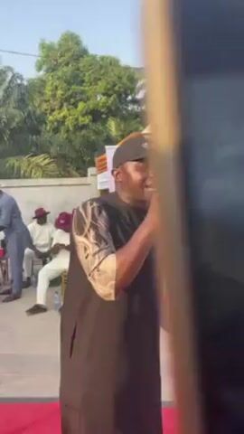 The video shows a man speaking into a microphone at an outdoor event. He is wearing a black cap, a dark sleeveless tunic over a patterned shirt. Behind him is a banner that reads "Nnokwa United Brothers". Several men are seated behind the banner, some wearing hats and traditional attire. One man is standing and appears to be serving them from a bag. The event appears to be held in a garden-like setting with trees in the background. The camera focuses on the man with the microphone as he speaks, occasionally panning to show the audience behind him.