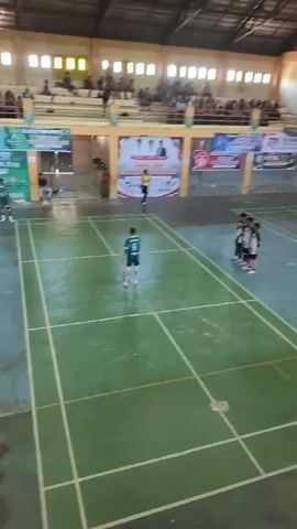 The video shows a futsal match taking place indoors in a gymnasium. Spectators are seated in tiered bleachers on the left side of the frame. A banner with text in a foreign language and images of people is displayed on the wall behind the court. The court is marked with white lines and appears to be made of a green, textured surface. On the court, there are several young people in athletic uniforms. One player in a green jersey with the number 5 on the back is standing in the center of the court with a futsal ball at his feet. Another player in a similar green jersey is on the left side of the court. On the right side of the court, a group of players in black and white striped jerseys stand in a line. A person in a yellow shirt, likely a referee, is standing near the center of the court, facing the player with the ball. The lighting in the gymnasium appears to be artificial, with some natural light possibly coming from windows at the top of the structure. The atmosphere seems to be one of a sporting event with participants and an audience.