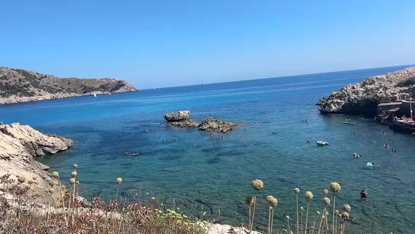 The video begins with a panoramic view of a clear blue ocean with a rocky coastline. Several people are seen swimming and enjoying the water. In the foreground, a few dried plants are visible. As the video pans to the right, a small establishment with umbrellas and tables is seen on a pier, with more people present. Beyond this, a parking lot filled with cars is visible, leading to a resort complex with multiple buildings on a hillside. The resort is predominantly white with blue accents and has numerous balconies. The sky is clear and bright blue. The camera continues to pan right, showing more of the resort and the surrounding landscape. The video ends with a view of the resort and the sea.