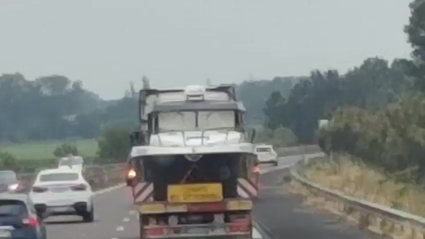 The video shows a rear view of a truck with a boat on its trailer on a highway. The truck is in the right lane of a multi-lane highway. The boat is white and appears to be a speed boat. There is a yellow sign on the back of the trailer with black text. Several cars are visible in the left lane and to the left of the truck. The highway appears to be in a rural or suburban area with trees and vegetation on the right side of the road. The sky is overcast. The video is shaky, indicating it was likely filmed from a moving vehicle.