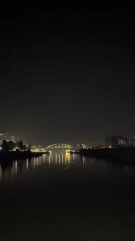 A vertical, handheld video captured at night shows a wide river with a bright, arched bridge in the distance. The bridge is illuminated with many lights, and its reflection is visible on the dark water. On either side of the river, there are city lights and some trees. The sky is dark, with a few faint stars visible. The camera appears to be stationary, or moving very slightly.