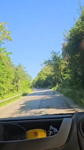 A first-person perspective video shows a car driving down a road lined with trees. The sky is clear and blue, and the sun is shining. The road is paved and appears to be in good condition. There are a few other cars visible on the road ahead. The trees are lush and green, and the overall impression is one of a peaceful drive through nature. In the foreground, a dash cam and a yellow object are visible.
