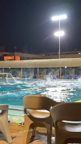 A water polo match is underway at night. Several players are in the water, actively swimming and splashing. One player appears to be in the process of shooting or has just shot the ball towards the goal. The goal net is visible in the background with a yellow ball in it. Bright stadium lights illuminate the pool. Spectator seating with chairs is in the foreground. The background shows a building with awnings and lights. Water is being sprayed into the air from the activity in the pool.