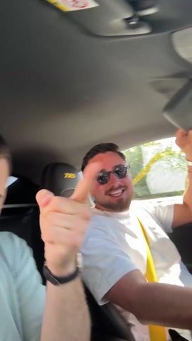 Two men are inside a car, listening to music. The driver is wearing sunglasses and a white t-shirt, with a yellow seatbelt. The passenger, wearing a light blue t-shirt and a bracelet, is dancing along to the music. The headrest of the seat reads "718." The video is shaky.