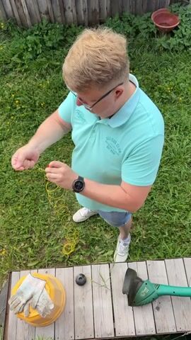 A person with blonde hair and glasses is standing in a grassy yard and is holding a yellow spool of string. The person is wearing a turquoise polo shirt, denim shorts, and white sneakers. A weed trimmer is visible on a wooden surface in front of the person, along with a yellow bucket and work gloves. The person is focused on the spool of string and appears to be preparing it for use.